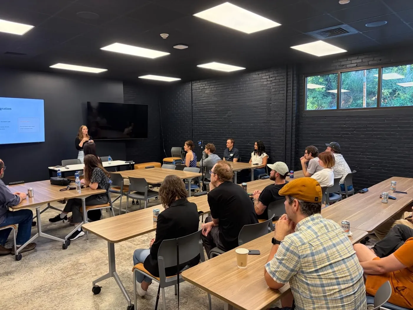An audience seated at long tables watching a presenter at the front of a dark brick room, at an AVL UX meetup